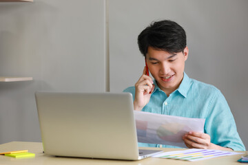 Young handsome male businessman with black short hair wearing light blue shirt working on office desk with silver laptop by looking at work document while using red mobile phone