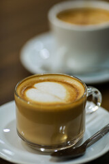 Close-up shot of white heart shape milk foam design and decorate on top of brown good smell coffee in clear glass cup on white ceramic dish with steel tea spoon serving on wooden table in cafe