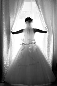 A bride in a white dress looks out the window. Black and white vertical photo. Rear view. Lens blur, film noise
