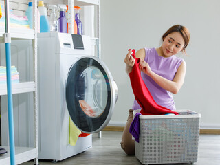 Young happy beautiful friendly asian female long brown ponytail hair housekeeper wear purple sleeveless shirt and brown pants put dirty clothes from basket into white washing machine in laundry room