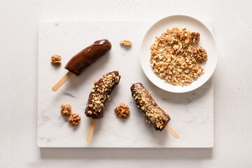 Frozen banana lollipops or lollies with walnuts sprinkles for kids on white. Easy vegan sweets. View from above.