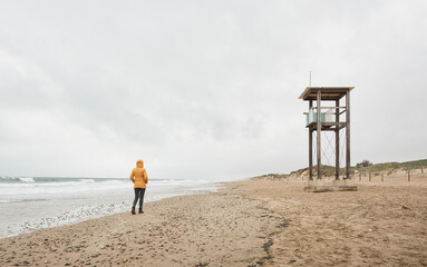 Girl in yellow raincoat looking at the sea. Walk along the beach on a bad weather day. Person walking on sand next to coast guard hut
