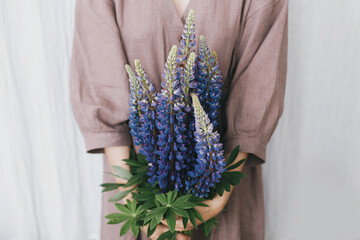 Stylish woman in linen dress holding lupine bouquet in hands closeup. Simple slow living. Aesthetics