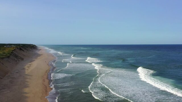 Aerial shot of people at beach by coastal town against blue sky, drone flying forward over waves splashing on shore - Cape Cod, Massachusetts