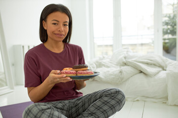 Happy young mixed race woman wearing casual clothes sitting on floor in cozy bedroom holding plate of colorful doughnuts, having breakfast. Food, dieting, bakery, calories and carbonhydrates