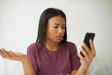Portrait of displeased young Afro American female posing isolated sitting on bed with cell phone,...
