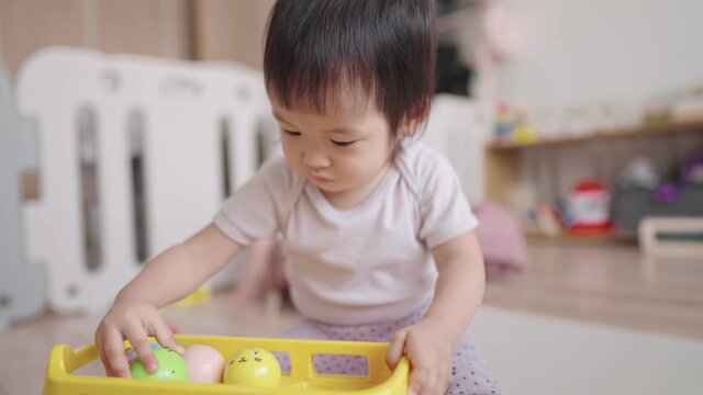 Smart active toddler picking up balls to put on ceiling of car toy in her hands, playful baby girl playing in house with being supervised by her private babysitter during a day while parent working