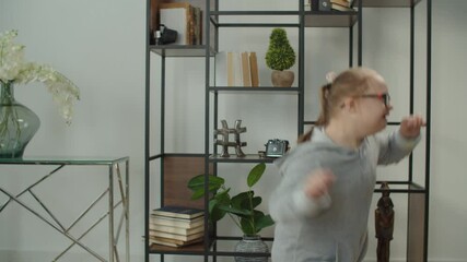 Playful mischievous cute preadolescent girl with general learning disability in eyeglasses having fun playing, jumping and spinning around in domestic room, expressing carefree mood and happiness.