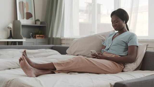 Wide shot of young African woman wearing wireless earphones sitting in bed at home with mobile phone in hands and listening to music
