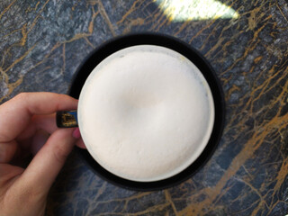 Hot drink coffee cup with capuccino and a foam on the top, top view, milk, white foam on a black plate on table with a hand holding a cup.