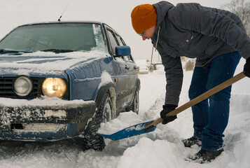 The guy is holding a shovel in his hands with snow near the car. Clearing a road for a car during a...