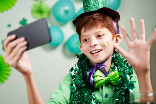 Young kid in green attire celebrating saint patricks day by making video call on mobile phone on decorated background during coronavirus covid-19 pandemic. - Powered by Adobe