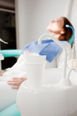 detail of dental chair with glass of water and tap, with a brunette female patient, in the background,