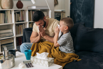Obraz premium father and son catch a cold. they are sitting in living room drinking tea and blowing their noses
