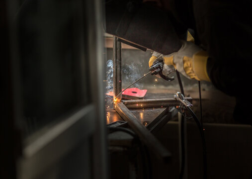 a welder in a welding mask, gloves welds metal with a welding machine and an electrode in his hand, sparks fly, a beautiful glow, bokeh, orange light on a black background, a flash of light and smoke