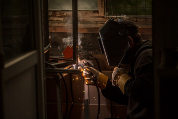 a welder in a welding mask, gloves welds metal with a welding machine and an electrode in his hand, sparks fly, a beautiful glow, bokeh, orange light on a black background, a flash of light and smoke