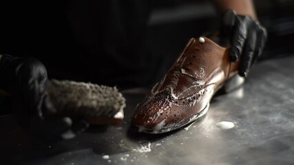 Close-up hands of shoemaker in black gloves applying cleaning foam on old light brown leather shoes. Process of repairing and restoration shoes by cobbler in shoe repair shop with dark interior.