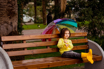 a beautiful little girl in yellow boots and a yellow sweater is sitting on a park bench with a colored umbrella