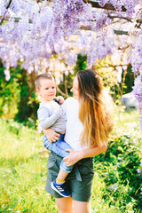Woman is holding her child in her arms under a blooming wysteria arms