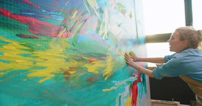 Close up of joyful happy Caucasian young female painter drawing with her hands on large canvas. Side view of contemporary artist creating colorful, sensual oil painting with her fingers in art studio