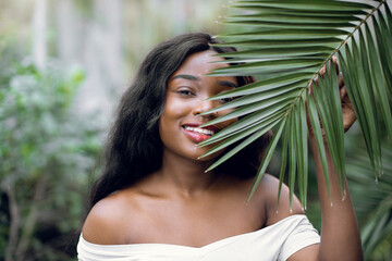 Fashion tropical portrait. Young attractive dark skinned woman model in white shirt, holding in hands green palm leaf while posing in beautiful greenhouse wth tropical plants.