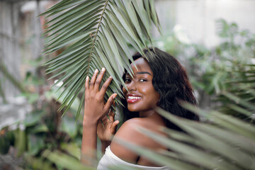 Young cute lovely African woman with perfect smooth skin, natural make up and long hair, smiling to camera, posing near tropical plants, touching palm leaf. Beauty and people concept.