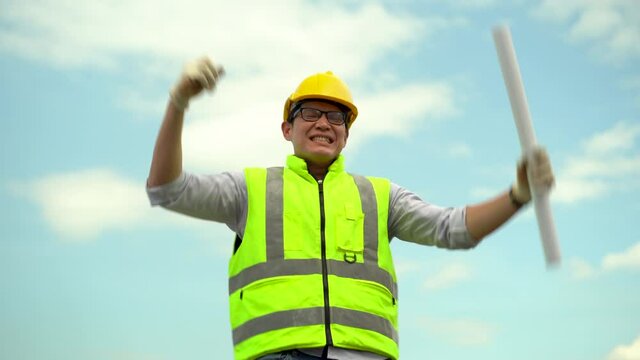 asian architect holding blueprint and fist up celebrating to succes  construction site with outdoors on blue sky . contractor worker or builder excited to finish work   Engineer or foreman glad  .