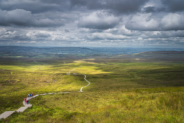 Fototapeta premium People hiking on wooden boardwalk winding between hills and fields, illuminated by sunlight, dramatic sky, Cuilcagh Mountain Park, Northern Ireland