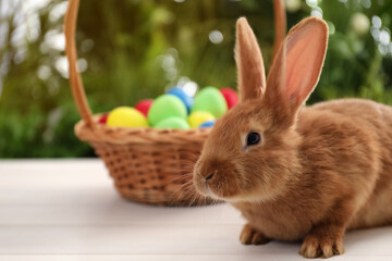 Cute bunny and basket with Easter eggs on table against blurred background. Space for text
