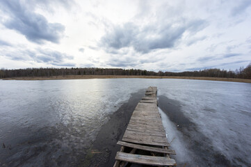Cold landscape of a frozen lake with a long wooden pier. Cloudy sky and forest on the far side of the pond.
