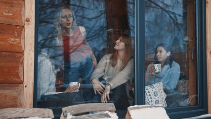Women, best friends spending winter vacation in wooden mountain house. Drinking hot beverages and gossiping near the big window. High quality photo