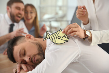 Young woman sticking paper fish to colleague's back while he sleeping in office, closeup. Funny joke