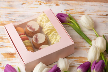 Pink gift box of desserts: macarons, shoux and zefyrs isolated on wooden background with white and purple tulips. Sweet present, beautiful flowers, flat lay. International women's day, birthday.