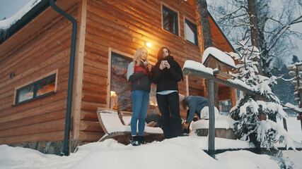 Friends spending winter holidays on the mountain. Drinking hot beverage in front of the wooden house and enjoying snowfall. High quality photo