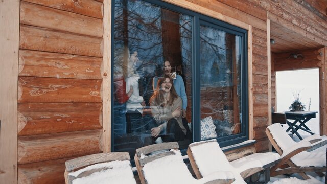 Beautiful woman best friends spending winter holidays in wooden mountain hut. Drinking hot beverage and looking through the window at snow. High quality photo