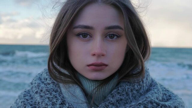 A portrait of a young woman in a sweater and a scarf against of a seashore. Seaside in autumn or winter, blue colors, windy weather