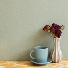 Gray mug cup and vase of dry rose flowers on wooden table. Khaki gray background