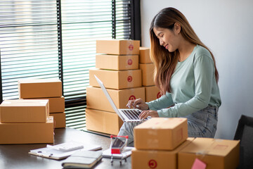 Young asian woman is entrepreneur work from home with online on notebook, online shopping, logistic and delivery, female is seller working on sofa, business SME