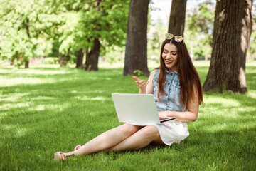 Cheerful girl communicating online using her laptop sitting on the lawn outdoors