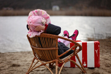 Cute 5-6 years old girl in a coat and summer hat sitting on a beach enjoying the beautiful view,...
