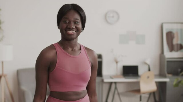 Lockdown portrait of young African-American sportswoman wearing trendy sportswear doing exercise with dumbbells in living room and looking at camera with smile on face