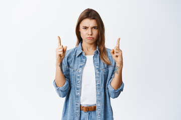 Thats bad. Upset young woman complain on unfair situation, frowning and sulking angry, pointing fingers up at disappointing offer, standing against white background