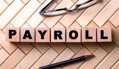 The word PAYROLL is written on wooden cubes on a wooden background next to a pen and glasses.