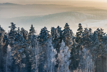 Winter landscape with forest, mountains, sky with clouds, frosty haze in the rays of the setting sun