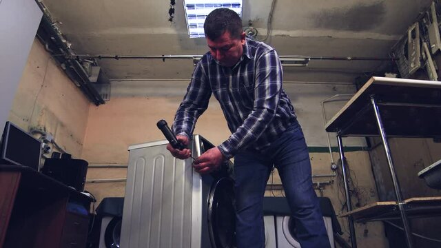 The man takes an electric screwdriver and unscrews the screw on the case. A home appliance technician fixes a breakdown in a workshop. Washing machine repair. The work of people in the service sector.