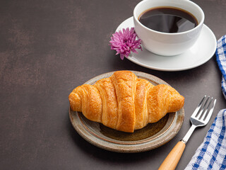 Top view of a croissant on a plate and a white coffee cup with a cloth and cutlery placed on a dark gray background. Space for text. Concept of relaxation