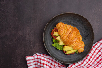 Top view of a croissant with avocados sliced, tomato, and lettuce on a black plate placed on a dark gray background. Space for text. Concept of foods