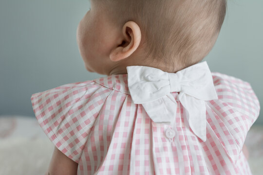 6 Month Old Baby Sitting Up Wearing A Pink Check Dress With White Bow; Girl Faces Away From Camera 