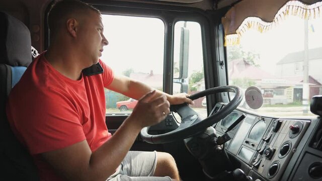 Male trucker holds hand on steering wheel and talks on mobile phone while driving a car. Lorry driver speaks on a smartphone during operating the truck through countryside. Close up Slow motion
