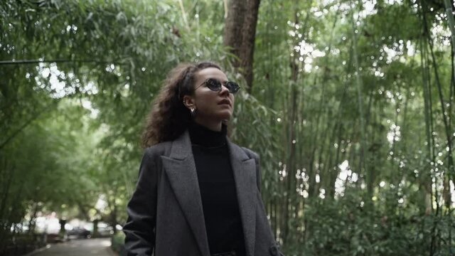 A young woman in sunglasses and a gray coat with a clutch walks in a green park. The sun's rays make their way through the bamboo thickets. Enjoy the fresh air.